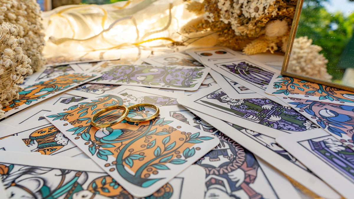 Fedi nuziali d'oro appoggiate su carte da gioco illustrate a mano, in controluce fra fiori secchi