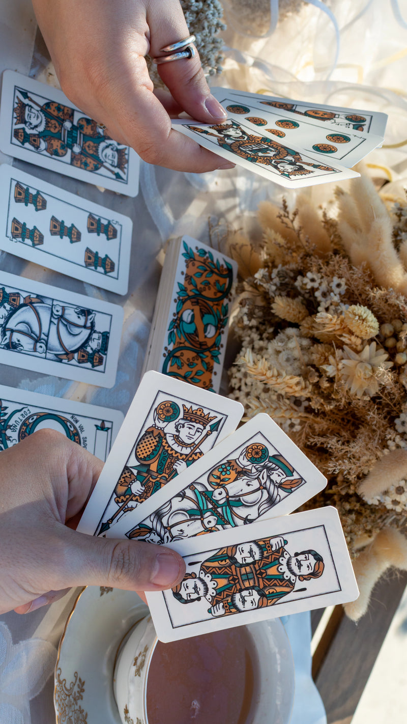 Due mani sfogliano un mazzo di carte turchesi fra i fiori secchi