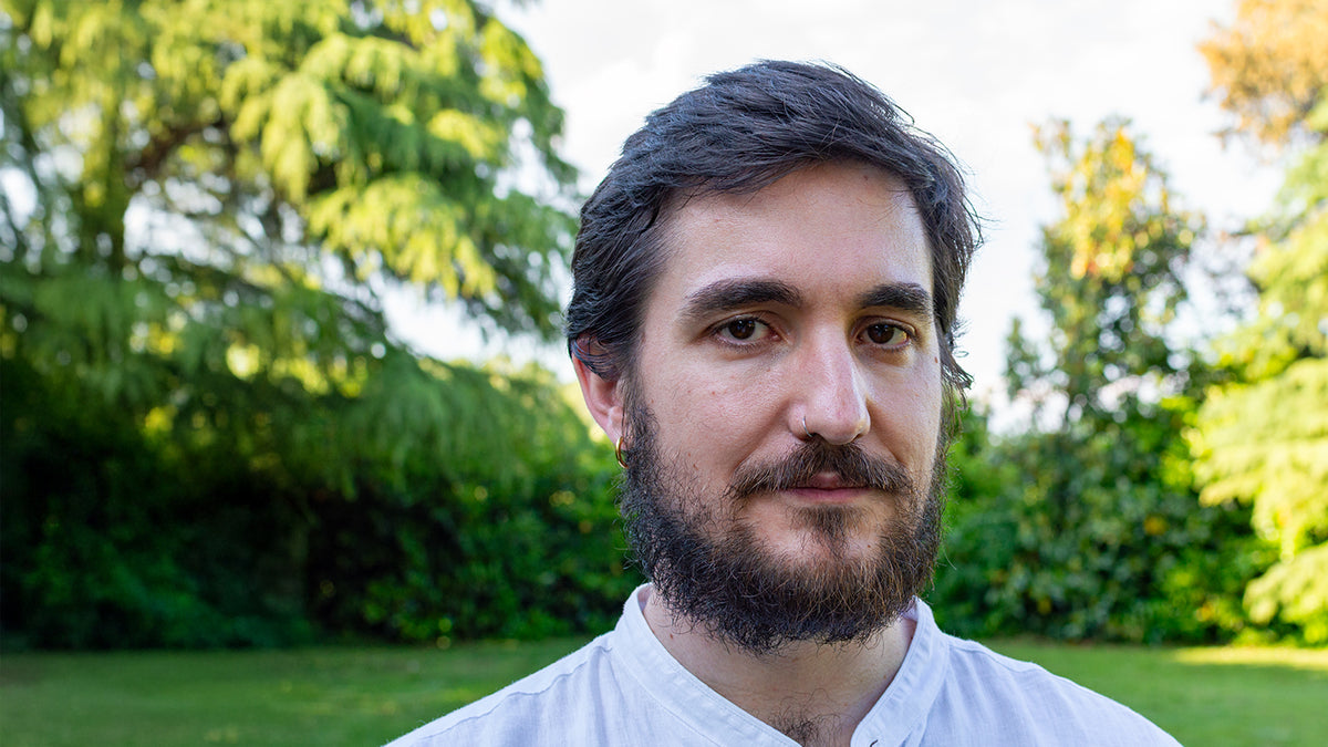 Ritratto fotografico di un uomo con barba scura, in camicia bianca, davanti al verde del giardino